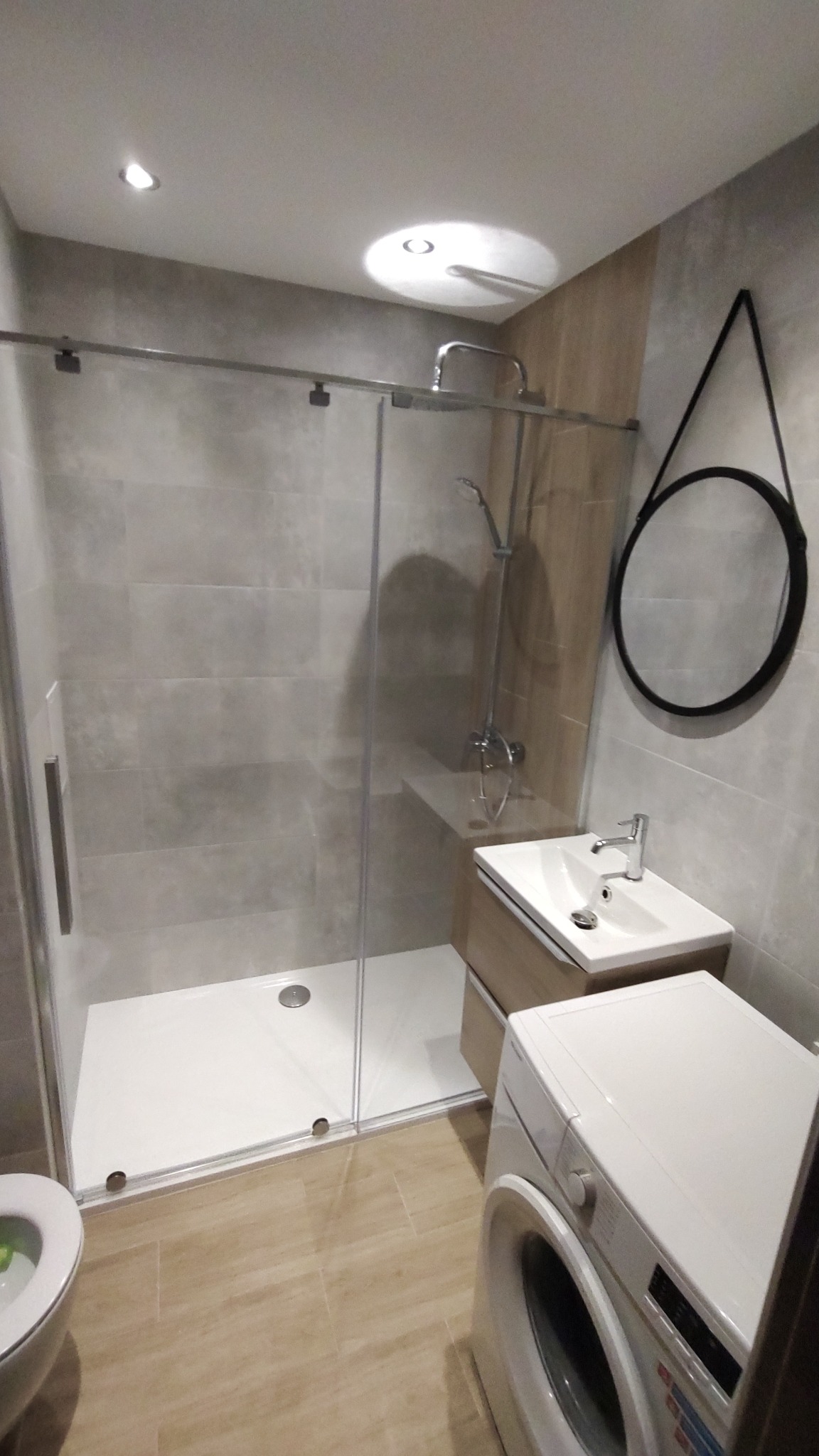 Nowoczesna łazienka z kabiną prysznicową typu walk-in, umywalką z szafką w kolorze drewna i okrągłym lustrem w czarnej ramie, pralka obok umywalki, szare płytki na ścianach i beżowe na podłodze.
