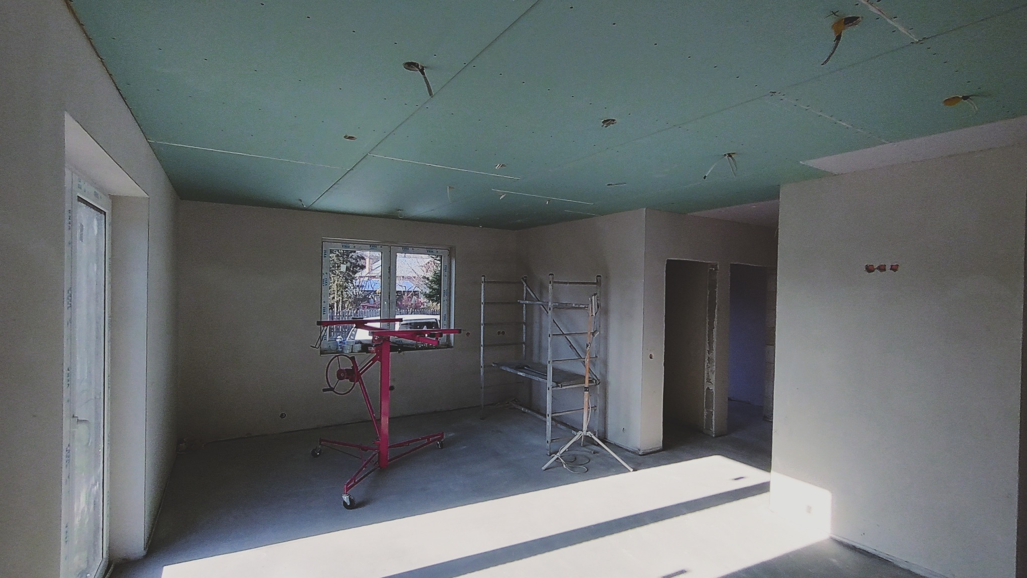 Wnętrze w trakcie prac wykończeniowych: ściany przygotowane pod gładź, sufit z płyt gipsowo-kartonowych, widoczny podnośnik do płyt oraz rusztowanie.