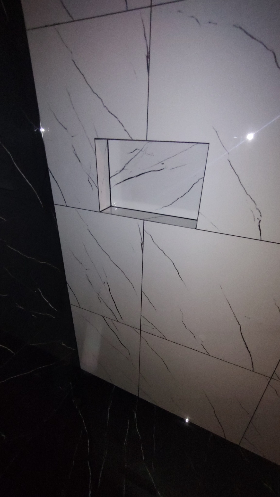 Wykończona ściana z białych płytek imitujących marmur z czarnymi żyłami, z wnęką na kosmetyki, obok czarnej ściany z podobnym wzorem.