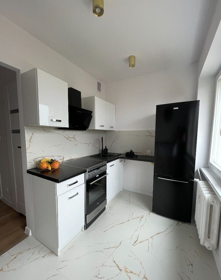 Nowoczesna, jasna kuchnia w Jeżowem z białymi szafkami z połyskiem, czarnym sprzętem AGD i marmurową podłogą z złotymi żyłami. Na blacie kosz z owocami.