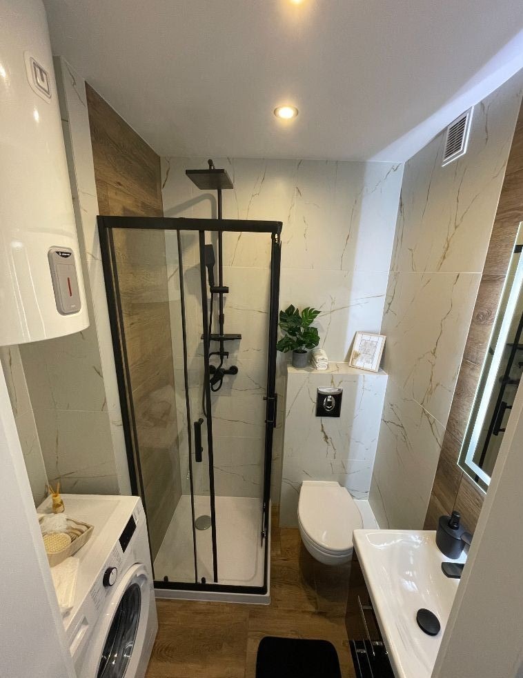 Nowoczesna łazienka z kabiną prysznicową w czarnej ramie, marmurowymi płytkami, drewnopodobną podłogą, pralką i minimalistyczną umywalką.