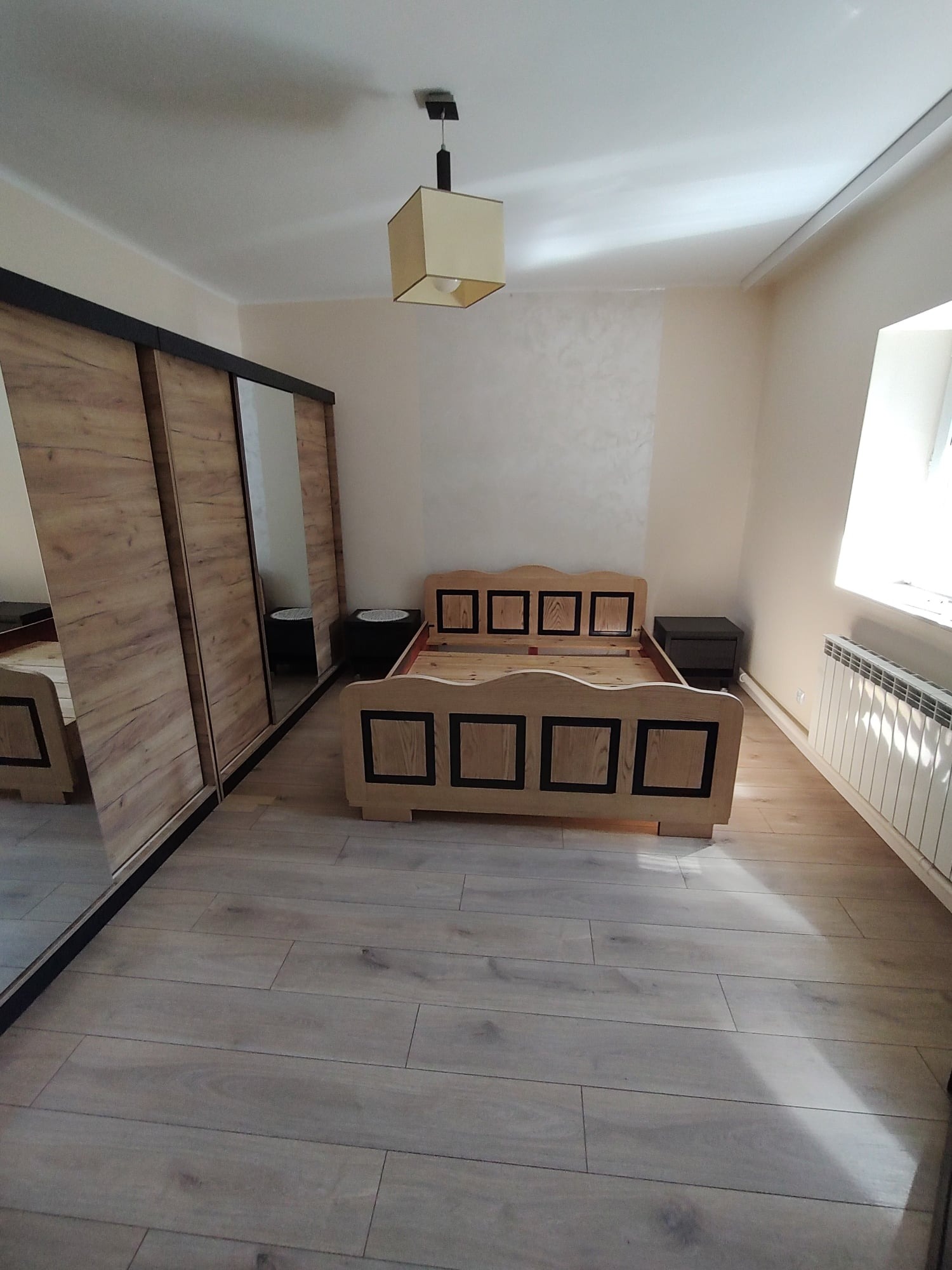 Sypialnia po remoncie: drewniana podłoga, łóżko z jasnego drewna z czarnymi akcentami, szafa przesuwna z lustrem, beżowe ściany z dekoracyjną fakturą, lampa sufitowa z beżowym abażurem.