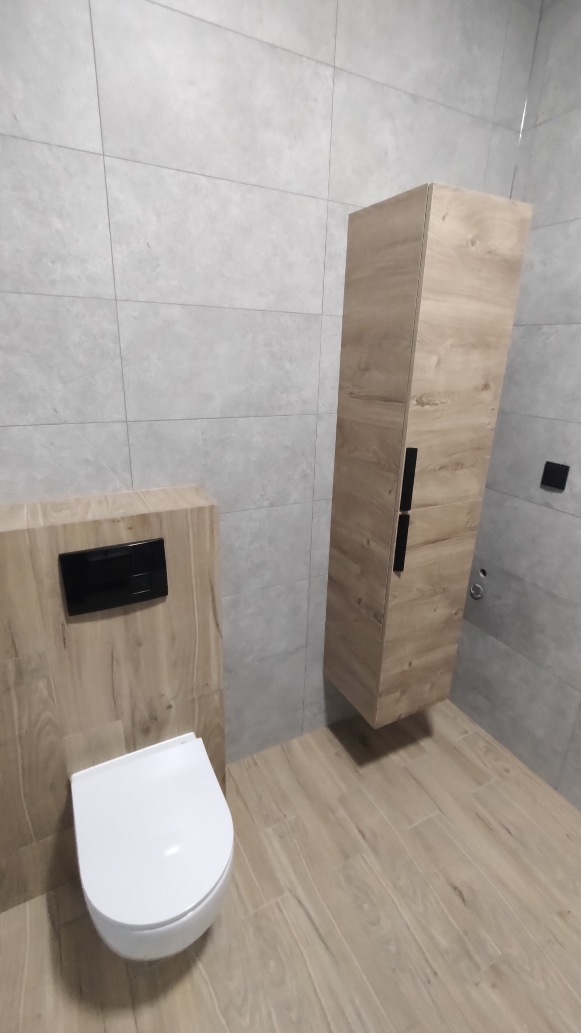 Nowoczesna toaleta podwieszana z drewnopodobną obudową, czarnym przyciskiem spłukującym i wisząca szafka łazienkowa z drewnianym frontem na tle szarych płytek.