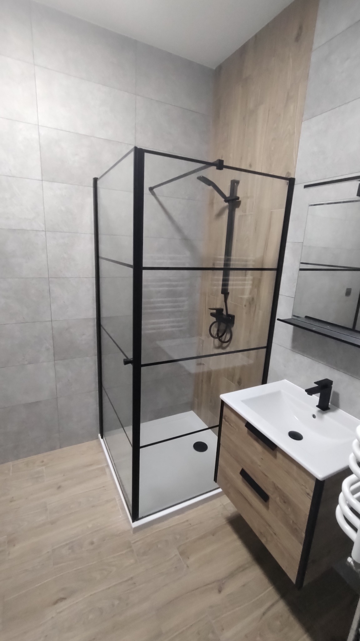 Nowoczesna łazienka z kabiną prysznicową typu walk-in z czarnymi profilami, umywalką nablatową z szafką w okleinie drewnopodobnej i czarną armaturą, wykończona płytkami w odcieniach szarości i drewna.