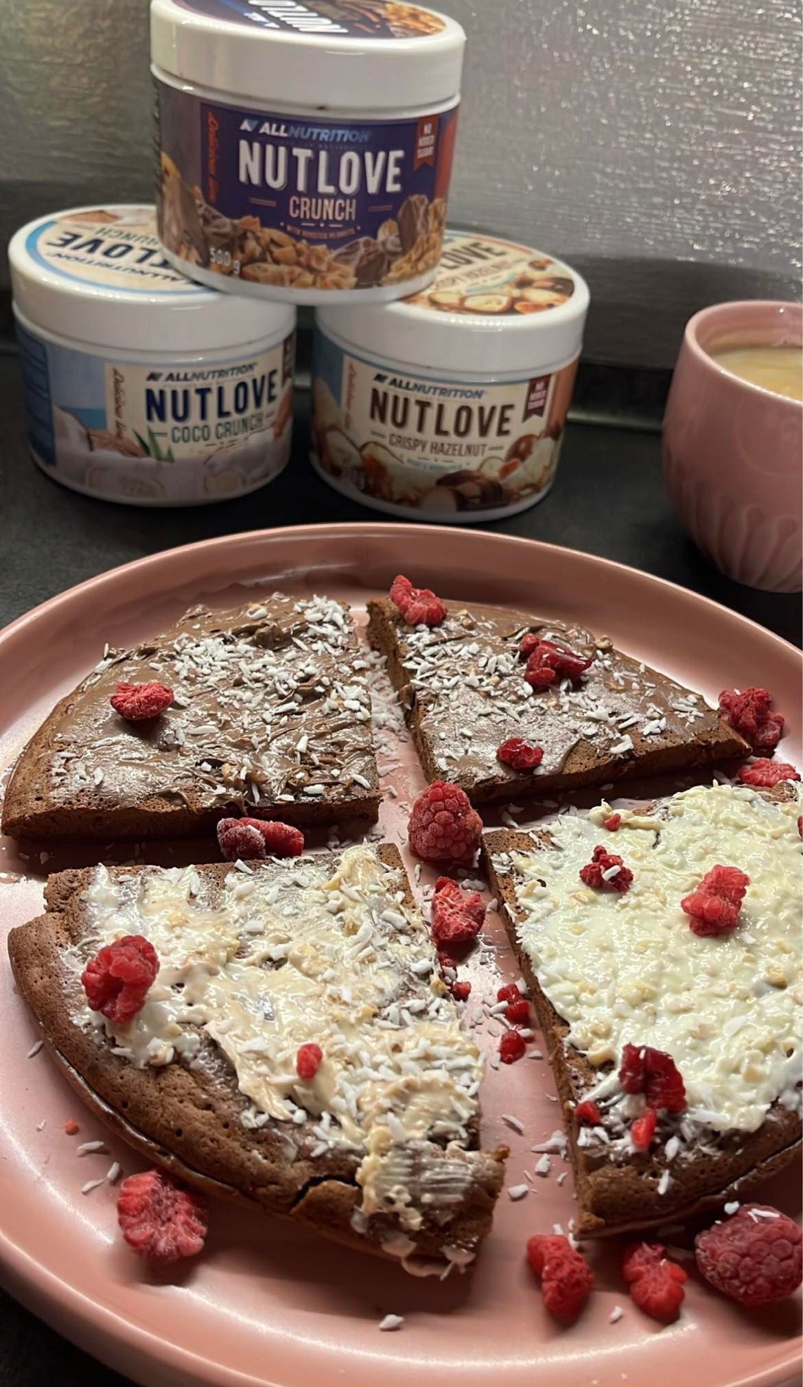 Cztery trójkątne kawałki ciasta proteinowego na różowym talerzu, posypane wiórkami kokosowymi i malinami, z różnymi smakami kremów, w tle trzy słoiki Nutlove i różowy kubek.