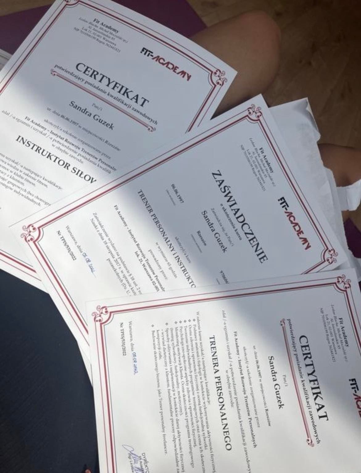 Stos certyfikatów i zaświadczeń Fit Academy dla Sandry Guzek, potwierdzających kwalifikacje instruktora siłowego i trenera personalnego.