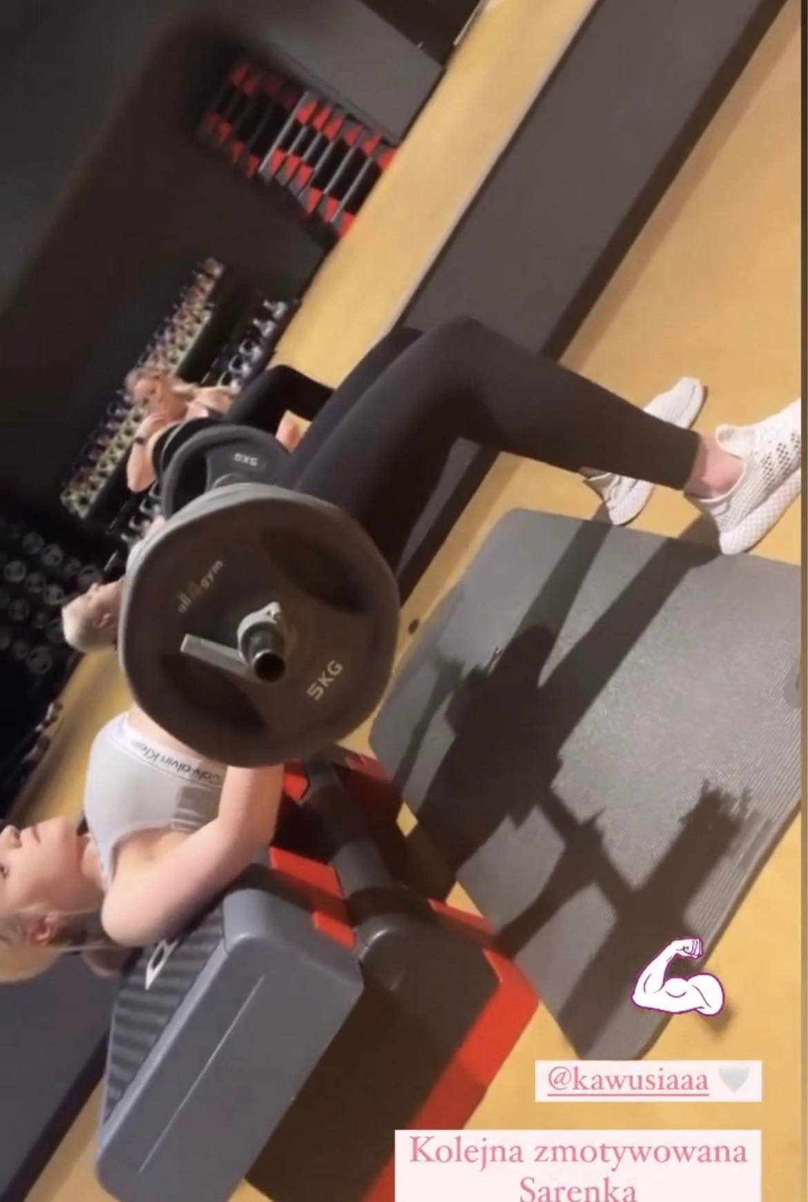 Dwie kobiety ćwiczą na siłowni, wykonując hip thrust z obciążeniem 5kg. Jedna w szarym topie, druga w czarnym, obie w czarnych legginsach i białych butach sportowych. W tle widoczne regały...