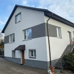 Dwukondygnacyjny dom po remoncie elewacji, z szarą dolną częścią i pasem na środku, brązowe rynny i dach, schody zewnętrzne z szarą balustradą, kostka brukowa przed domem.