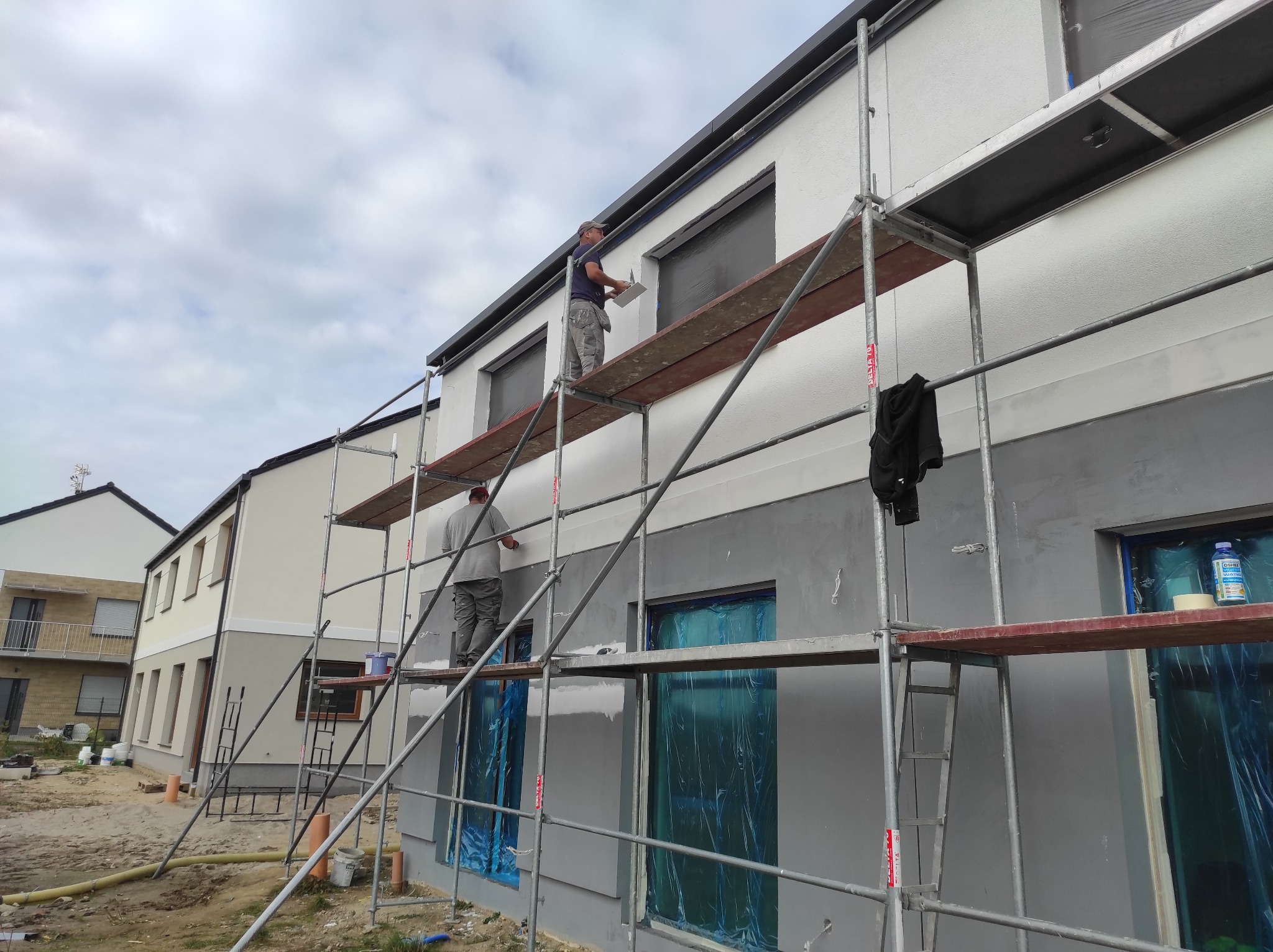 Dwaj pracownicy na rusztowaniu podczas nakładania białego tynku na szarą elewację budynku. Okna zabezpieczone folią. Widoczne narzędzia i materiały budowlane.