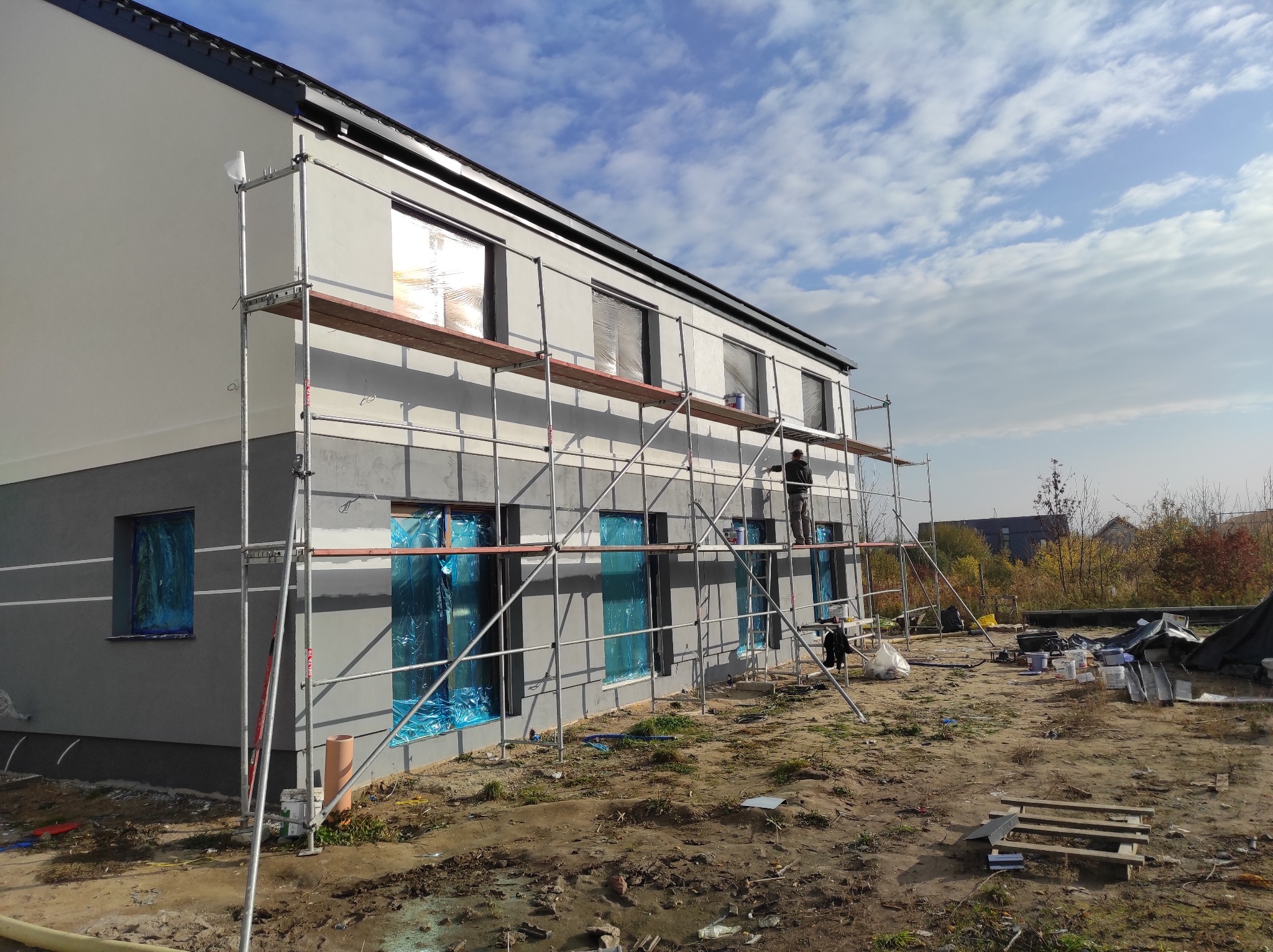 Dwukondygnacyjny dom w trakcie malowania elewacji, okna zabezpieczone folią, pracownik na rusztowaniu, widoczne farby i narzędzia.