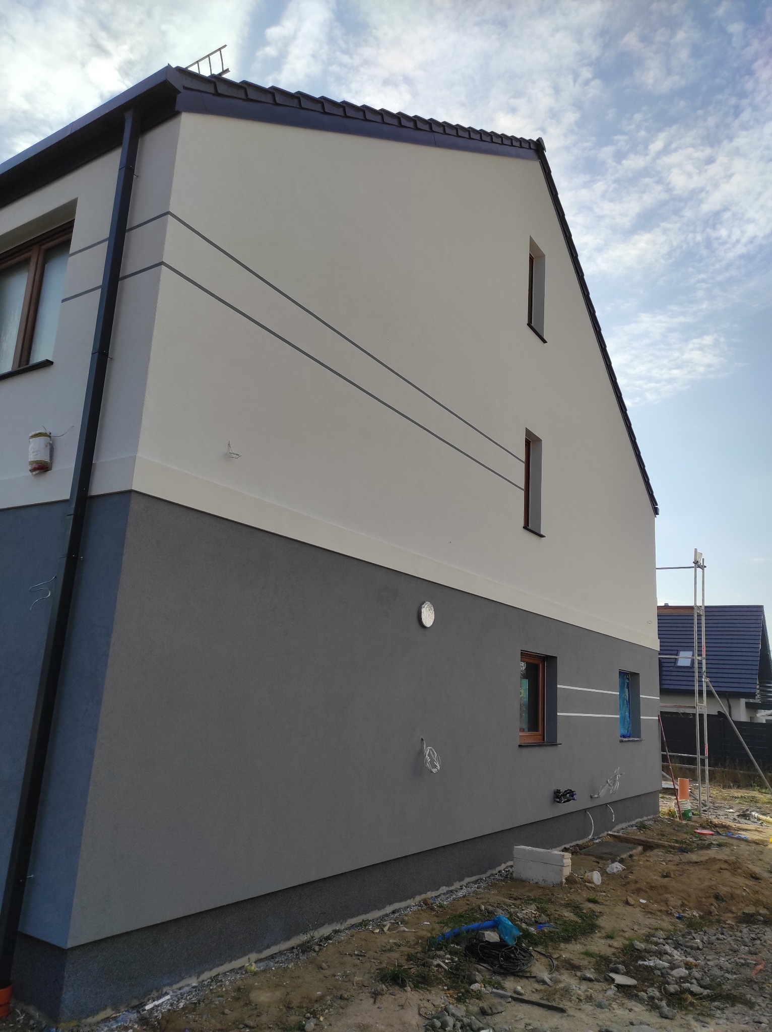 Dwukolorowa elewacja domu jednorodzinnego z szarymi pasami dekoracyjnymi, widoczne rusztowanie i elementy instalacji zewnętrznej.