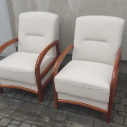 Dwa kremowe fotele z drewnianymi podłokietnikami, ustawione na brukowanym chodniku przed szarą ścianą. Fotele mają proste, eleganckie linie i wyglądają na świeżo odnowione.