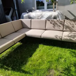 Beżowa, modułowa sofa tapicerowana z metalową ramą, ustawiona na trawie w ogrodzie, z widocznymi cieniami drzew i fragmentem budynku w tle.