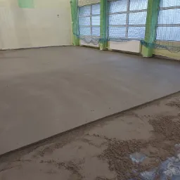 Świeżo wylana, gładka posadzka jastrychowa w hali sportowej, widoczne okna z siatką zabezpieczającą i częściowo rozłożona folia ochronna przy krawędzi.
