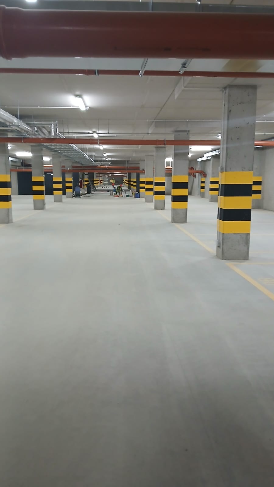 Wnętrze parkingu podziemnego z pomalowanymi na żółto-czarno kolumnami. W tle widoczni pracownicy podczas wykonywania prac wykończeniowych.