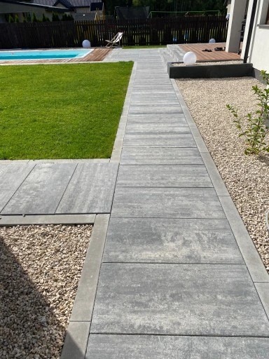 Nowoczesna ścieżka z szarych płyt brukowych prowadząca przez trawnik w kierunku basenu i drewnianego tarasu. Jasne kamienie dekoracyjne po bokach.