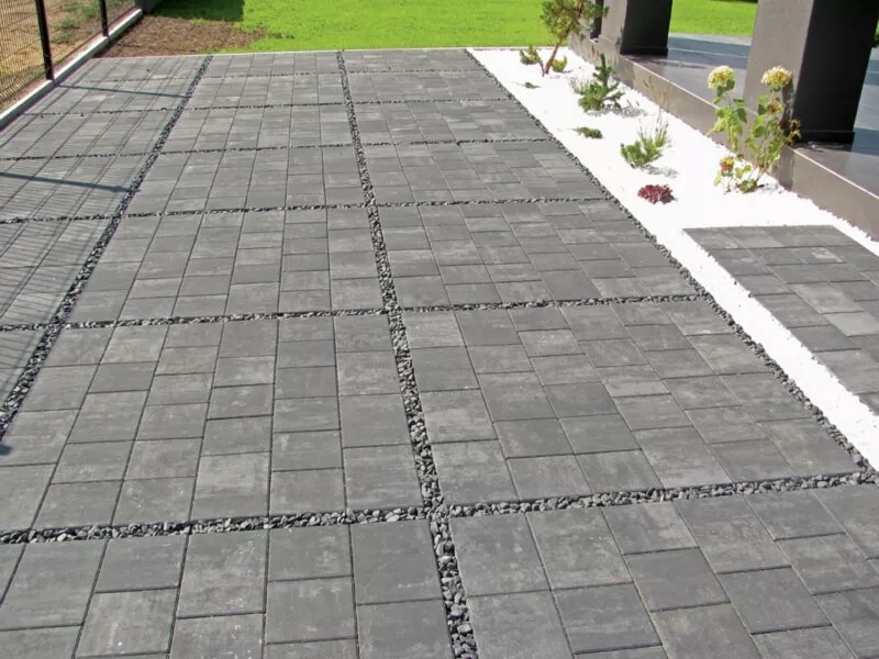 Szara kostka brukowa ułożona w kwadraty, oddzielona drobnymi kamieniami. Białe kamienie i rośliny przy budynku w tle. Estetyczne wykończenie podjazdu.
