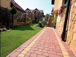 Kaskada kostki brukowej w odcieniach czerwieni i żółci, wijąca się przez zadbany trawnik wzdłuż kamiennej ściany domu. Schody prowadzą na górę, a kwiaty dodają koloru.