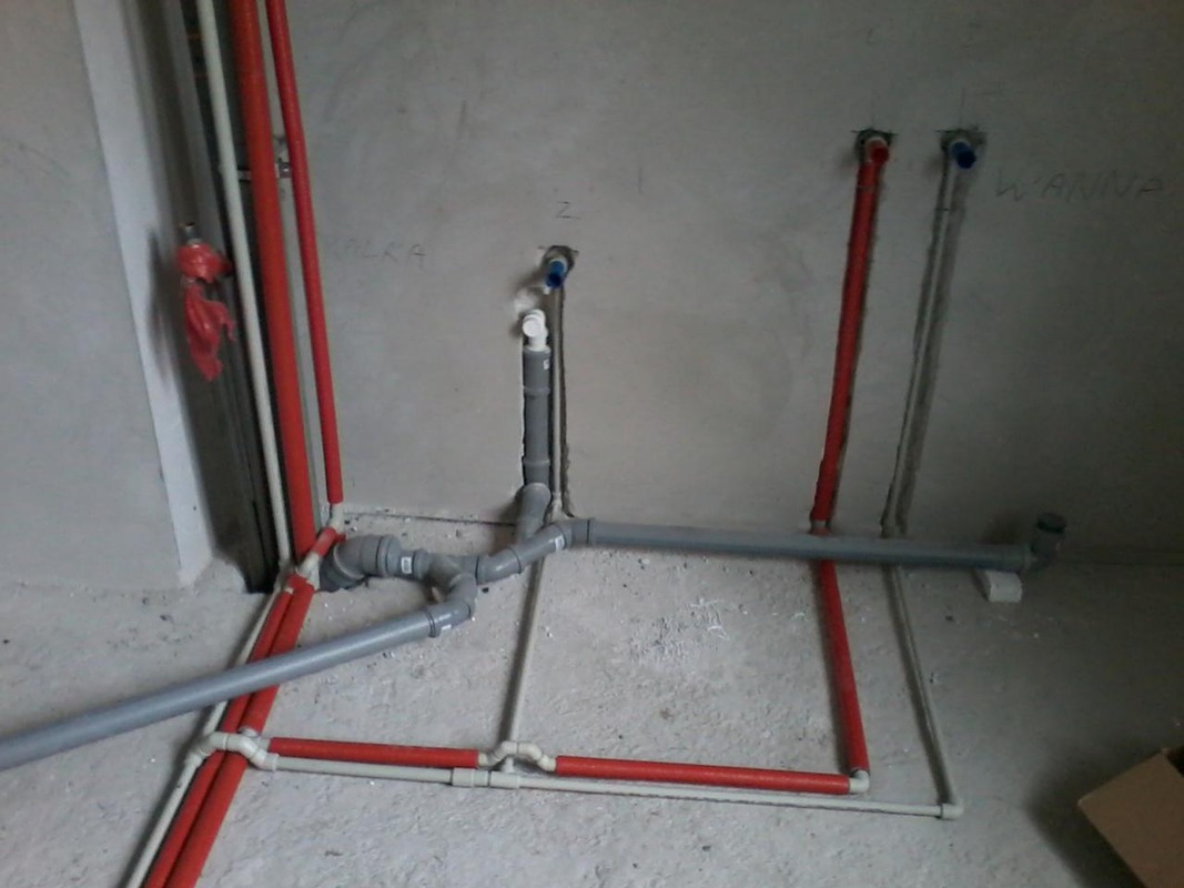 Instalacja wodno-kanalizacyjna w surowym stanie: czerwone i szare rury prowadzone po podłodze i ścianach, przygotowane do podłączenia urządzeń sanitarnych.