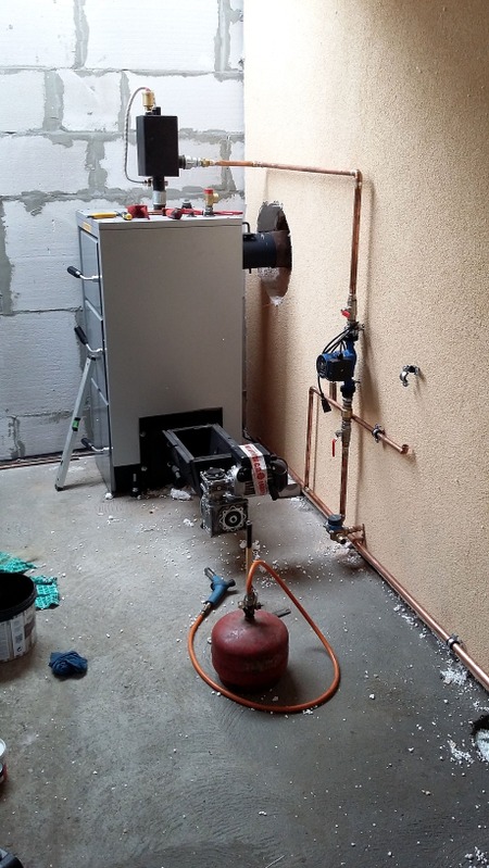 Montaż kotła grzewczego z podłączeniem miedzianych rur w surowym pomieszczeniu. Widoczne narzędzia i resztki materiałów budowlanych na podłodze.