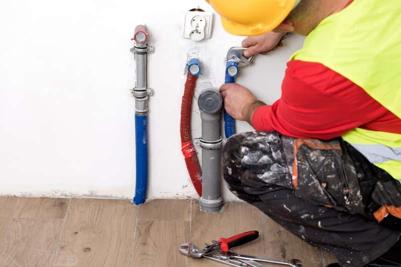 Instalator w kasku łączy rury przy ścianie, używając klucza nastawnego. Widoczne narzędzia na podłodze i fragment instalacji z kolorowymi rurami.