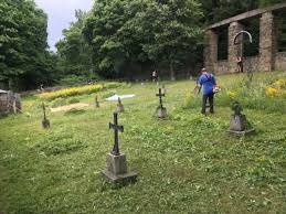 Kosiarka spalinowa w akcji na cmentarzu: mężczyzna w niebieskiej koszulce kosi trawę między nagrobkami, w tle zieleń drzew i ruiny kaplicy.