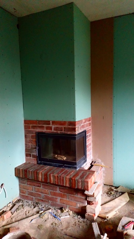 Wnętrze w trakcie budowy kominka z cegły, z widocznymi płytami gipsowo-kartonowymi na ścianach. Surowy stan wykończenia, widoczne materiały budowlane.