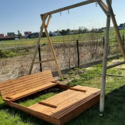 Drewniana konstrukcja huśtawki i piaskownicy w trakcie montażu na trawniku, widoczne elementy składowe i niekompletna piaskownica z pokrywą, w tle ogrodzenie panelowe i zabudowania.