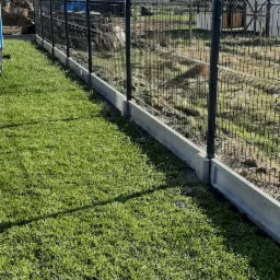 Nowoczesne ogrodzenie panelowe z podmurówką betonową na tle trawnika i budynków w trakcie budowy.