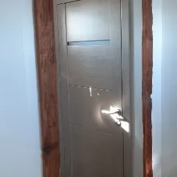 Lustro w drewnianej ramie z żarówkami dekoracyjnymi, odbijające drzwi wewnętrzne z poziomymi panelami i klamką, na tle białej ściany i drewnianej podłogi.