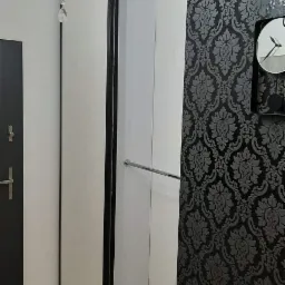 Biała szafa wnękowa z przesuwnymi drzwiami, widoczna w korytarzu z szaro-czarną tapetą w ornamenty i ozdobnym żyrandolem.