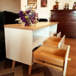 Stolik kawowy z białymi bokami, drewnianym blatem i wysuwanymi szufladami, na blacie bukiet kwiatów, w tle komoda i zdjęcia na ścianie.