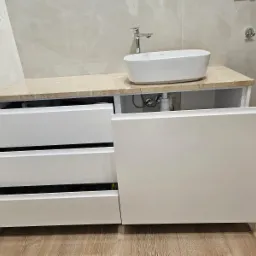 Nowoczesna łazienka z białą, owalną umywalką nablatową i szafką z trzema szufladami, z których jedna jest otwarta, ukazując zawartość. Blat szafki imituje marmur. Na ścianie widoczne wieszaki...