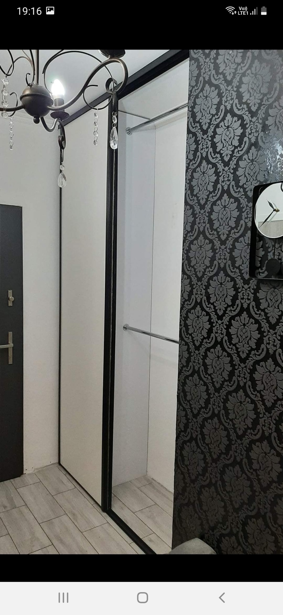 Biała szafa wnękowa z przesuwnymi drzwiami, widoczna w korytarzu z szaro-czarną tapetą w ornamenty i ozdobnym żyrandolem.