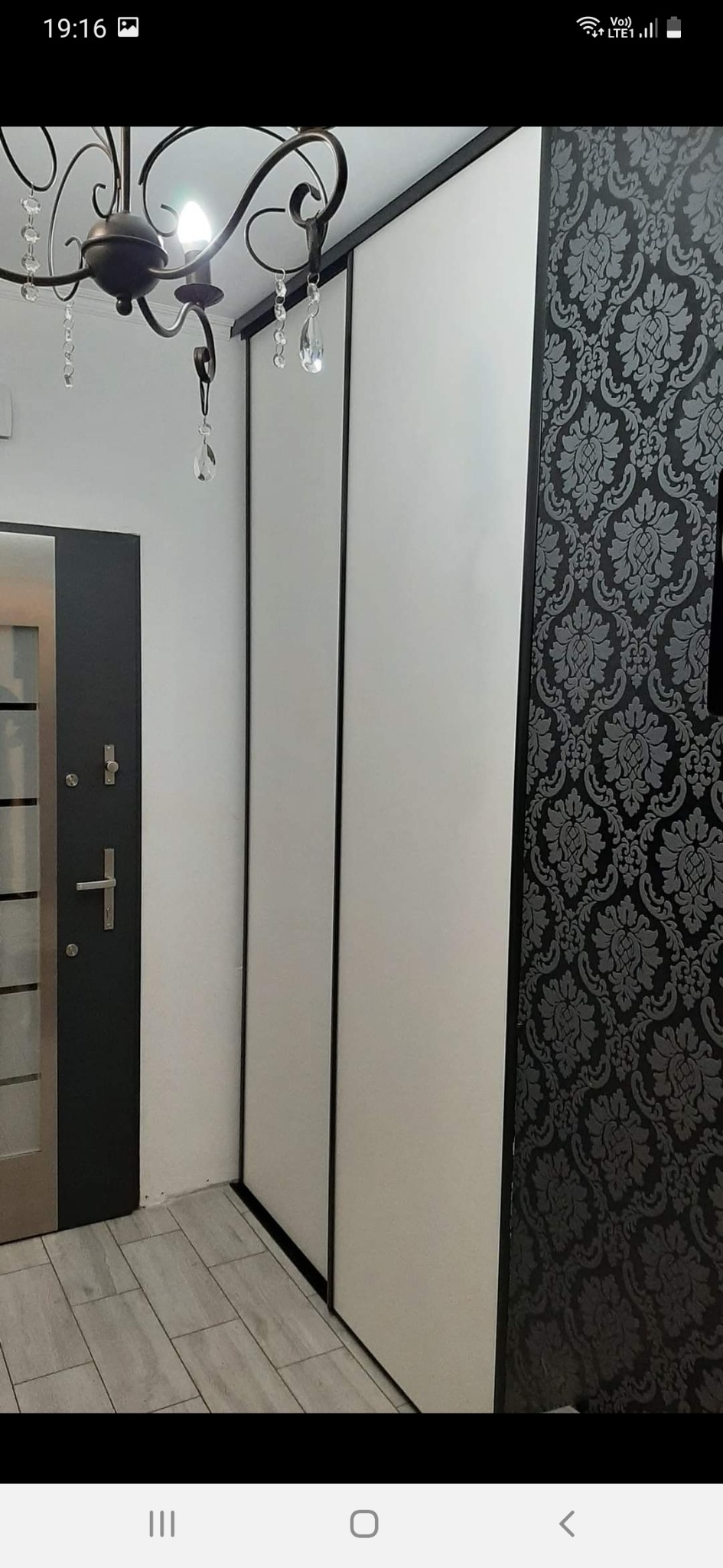 Biała szafa wnękowa z przesuwnymi drzwiami na tle ściany z czarno-szarym wzorem, widoczny fragment podłogi z płytek imitujących drewno, drzwi wejściowe i żyrandol.