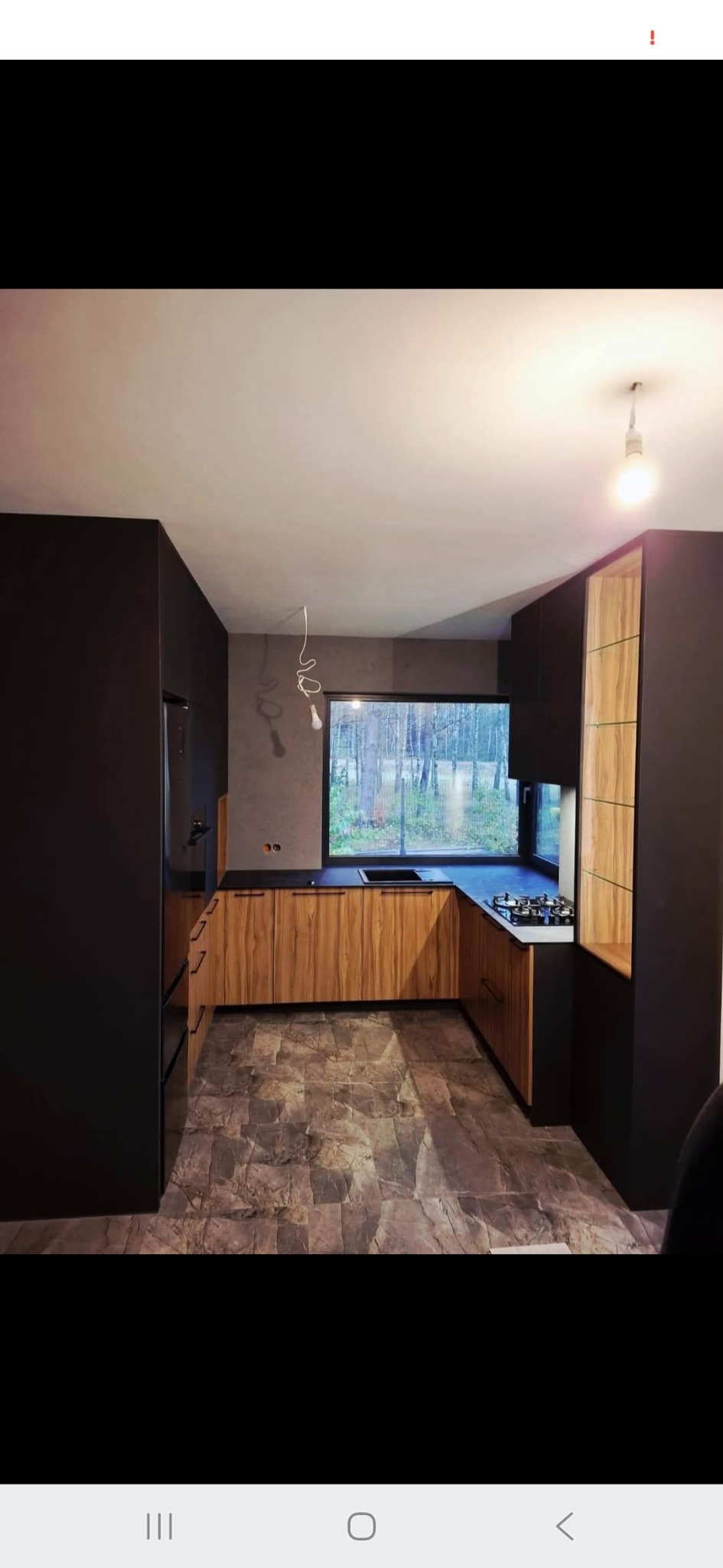 Nowoczesna kuchnia w odcieniach brązu i czerni z dużym oknem. Drewniane fronty szafek kontrastują z ciemnymi ścianami i blatem. Na podłodze płytki imitujące kamień.