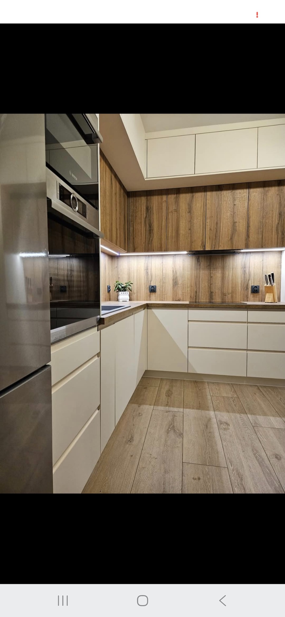 Nowoczesna kuchnia z drewnianymi akcentami: beżowe szafki dolne, drewnopodobne górne, oświetlenie LED podszafkowe, sprzęt AGD marki Bosch i podłoga z paneli.