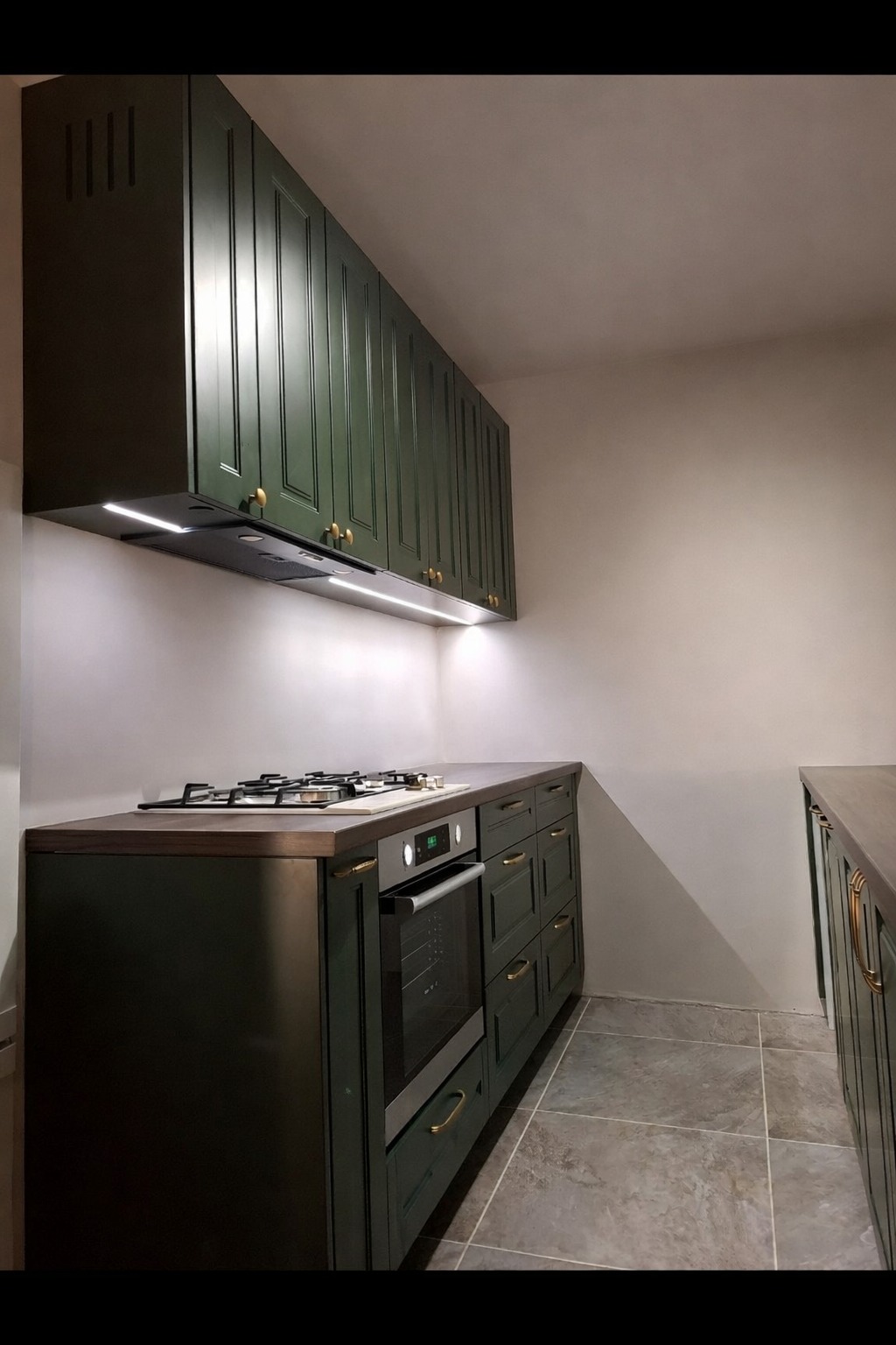 Nowoczesna kuchnia w odcieniach zieleni z złotymi uchwytami, oświetleniem LED pod szafkami wiszącymi i płytkami podłogowymi w szarym odcieniu. Widok na zabudowę kuchenną z piekarnikiem.
