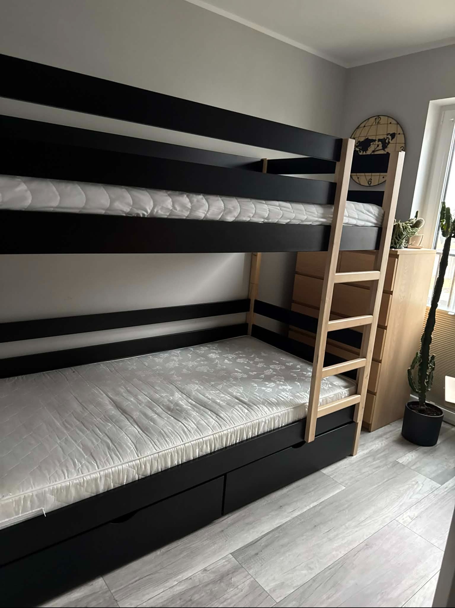 Dwupiętrowe łóżko z czarną ramą i drewnianą drabinką w pokoju z szarą podłogą i ścianami. Obok komoda i kaktus w doniczce. Na ścianie zegar z mapą świata.