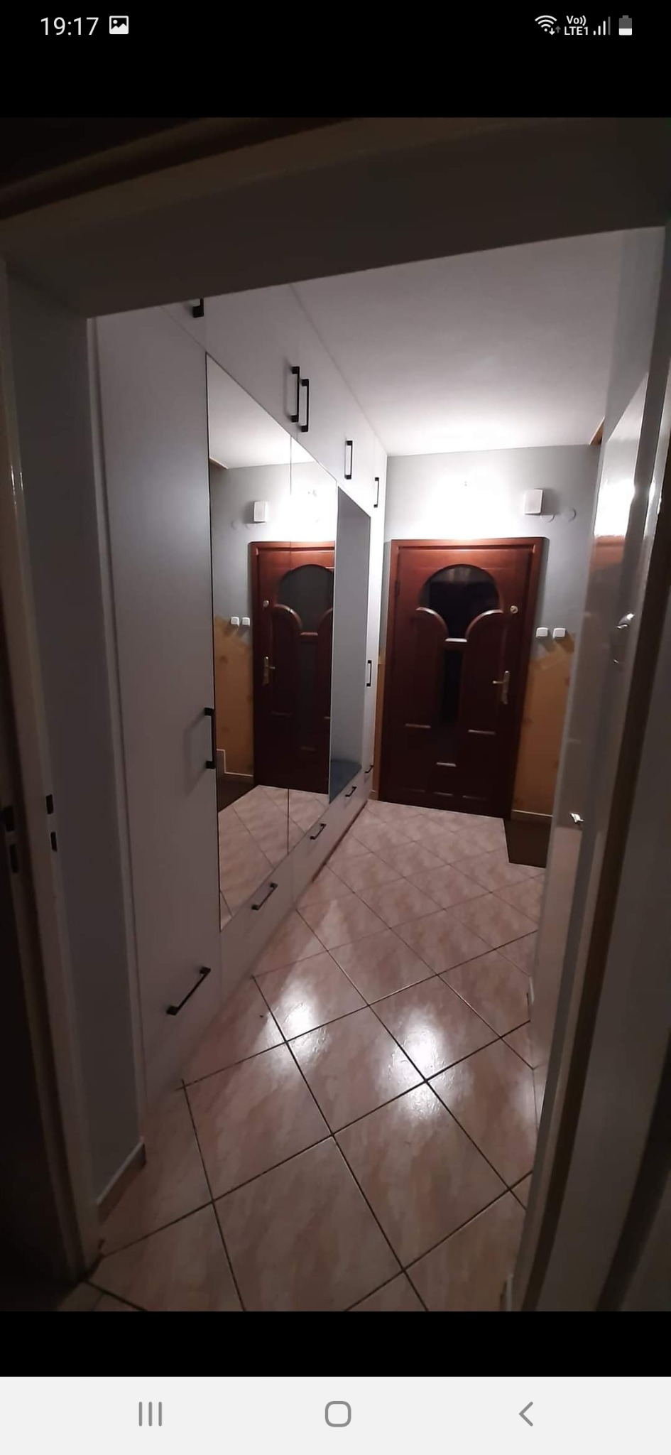 Wnętrze przedpokoju z dużą, białą szafą z lustrami na frontach i długimi, czarnymi uchwytami, widok na brązowe drzwi wejściowe z dekoracyjnym przeszkleniem.