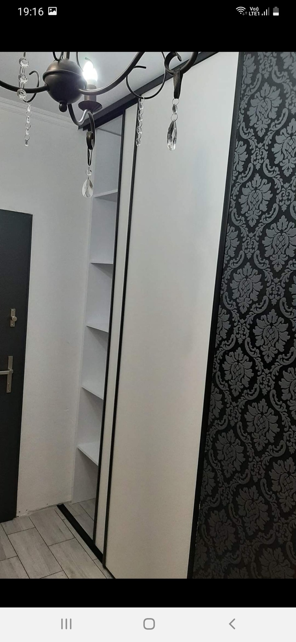 Biała szafa przesuwna z czarnymi obramowaniami, z otwartą boczną wnęką z półkami, na tle wzorzystej tapety w odcieniach szarości i czerni, oświetlona ozdobnym żyrandolem z kryształkami.