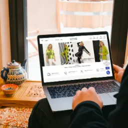 Osoba przegląda na laptopie stronę sklepu internetowego z odzieżą damską, widoczny czajnik i miseczka na ozdobnej podstawce, naturalne światło wpadające przez okno.