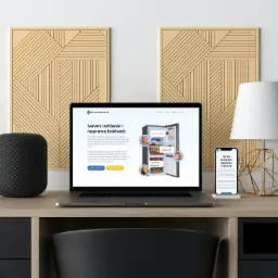 Prezentacja strony internetowej serwisu lodówek na laptopie i smartfonie, stojących na biurku obok głośnika, z dekoracyjnymi panelami na ścianie w tle i lampą ze złotym geometrycznym stelażem.