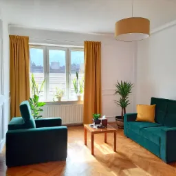 Jasny pokój z zielonym, welurowym zestawem wypoczynkowym, drewnianym stolikiem kawowym i roślinami doniczkowymi na parapecie i obok sofy, w tle białe ściany z dekoracyjnymi panelami i parkiet...