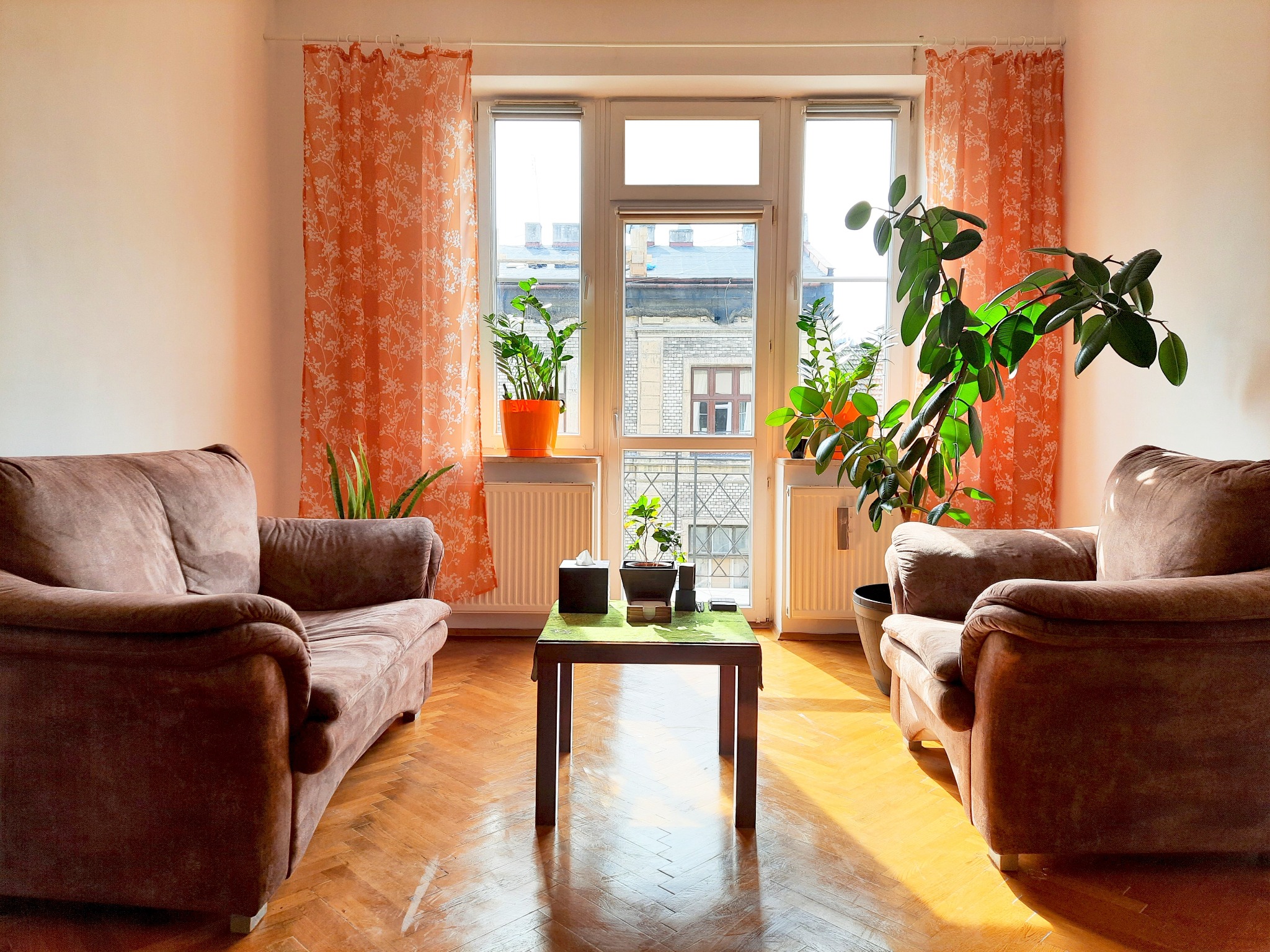 Jasny salon z dwoma brązowymi sofami, drewnianym parkietem, stołem nakrytym zielonym obrusem i roślinami doniczkowymi przy oknie z pomarańczowymi zasłonami.