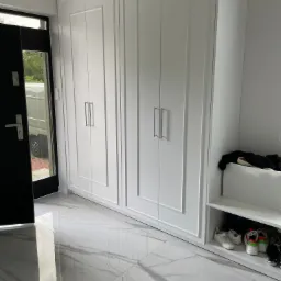 Biała szafa wnękowa z dwoma drzwiami, zintegrowana z półką na buty, na podłodze marmurowe płytki z wyraźnym żyłowaniem, widok na otwarte drzwi wejściowe i zaparkowany samochód dostawczy.