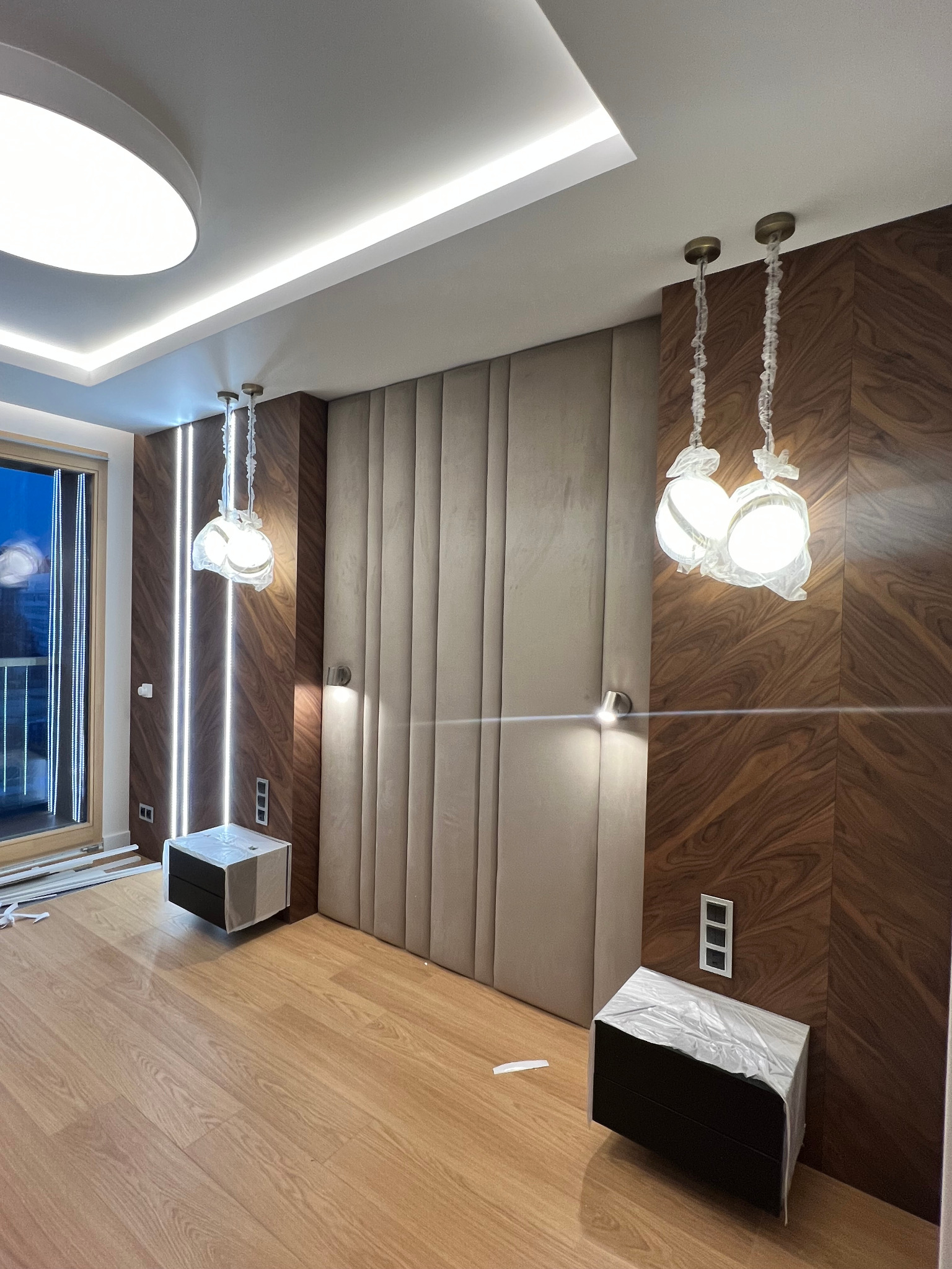 Wnętrze sypialni w trakcie wykończenia: drewniana podłoga, ściana z tapicerowanymi panelami, drewniana zabudowa z oświetleniem LED, szafki nocne w folii ochronnej, wiszące lampy w osłonach.