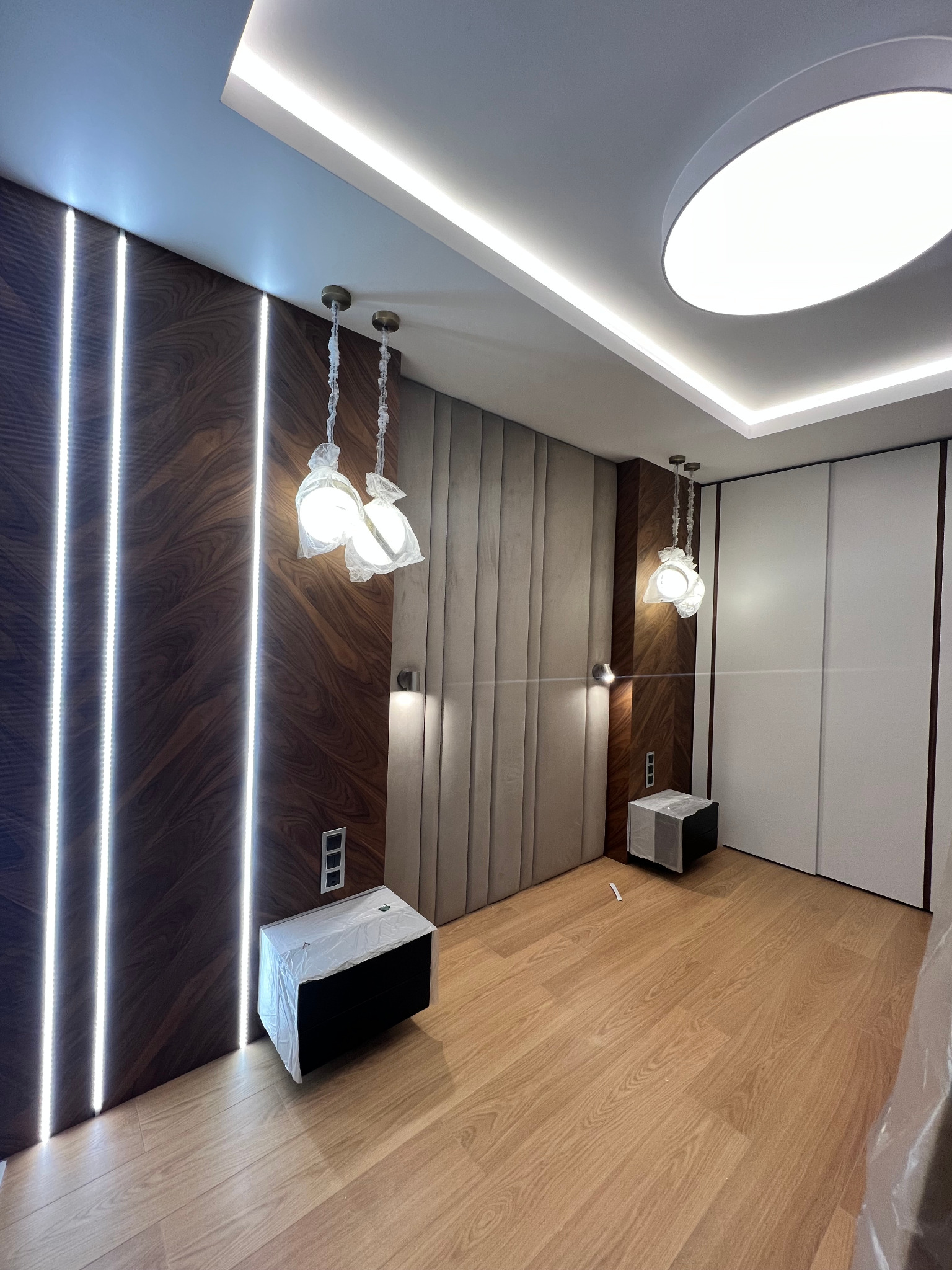 Nowoczesna sypialnia z drewnianą ścianą z pionowymi paskami LED, tapicerowanym panelem za łóżkiem, wiszącymi lampami w folii ochronnej i szafami wnękowymi po prawej stronie.