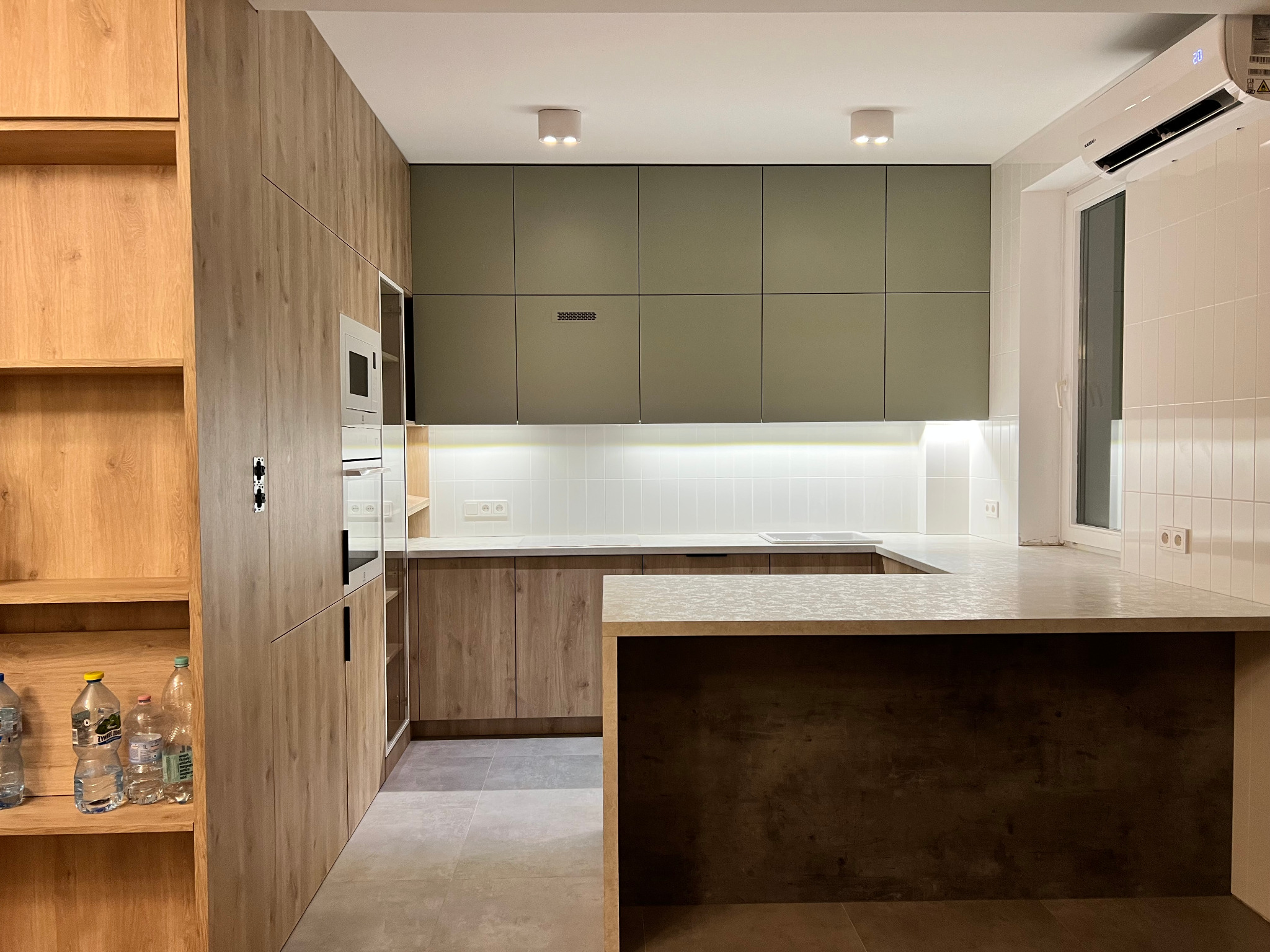 Nowoczesna kuchnia z drewnianymi frontami szafek, oliwkowymi górnymi szafkami, wyspą z ciemnym wykończeniem i białymi płytkami na ścianie, oświetlona punktowymi lampami i klimatyzatorem nad oknem.
