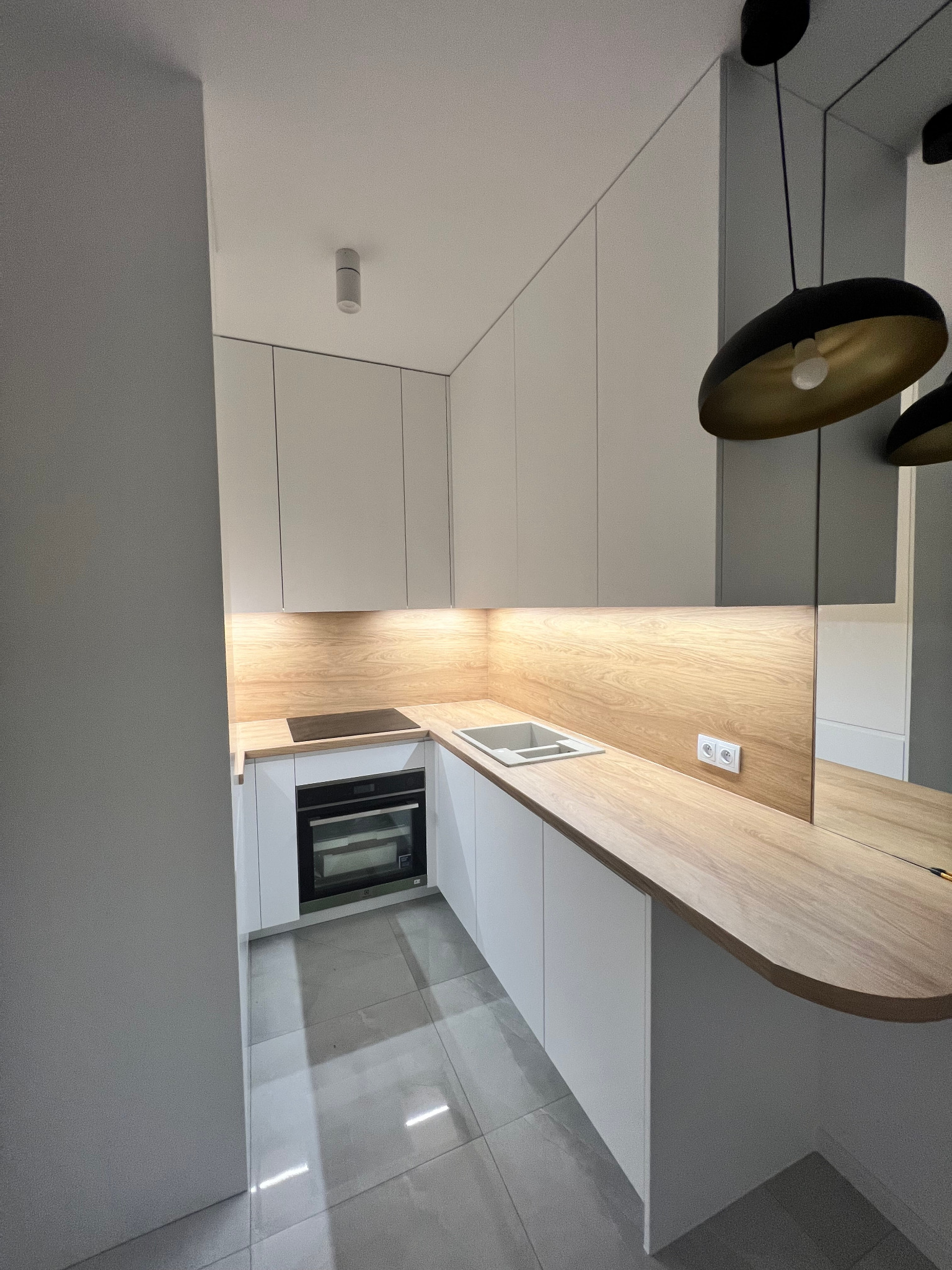 Nowoczesna, minimalistyczna kuchnia z białymi szafkami, drewnianym blatem i panelem ściennym, oświetlona od dołu, z czarnymi wiszącymi lampami z elementami w kolorze złota i szarymi płytkami...
